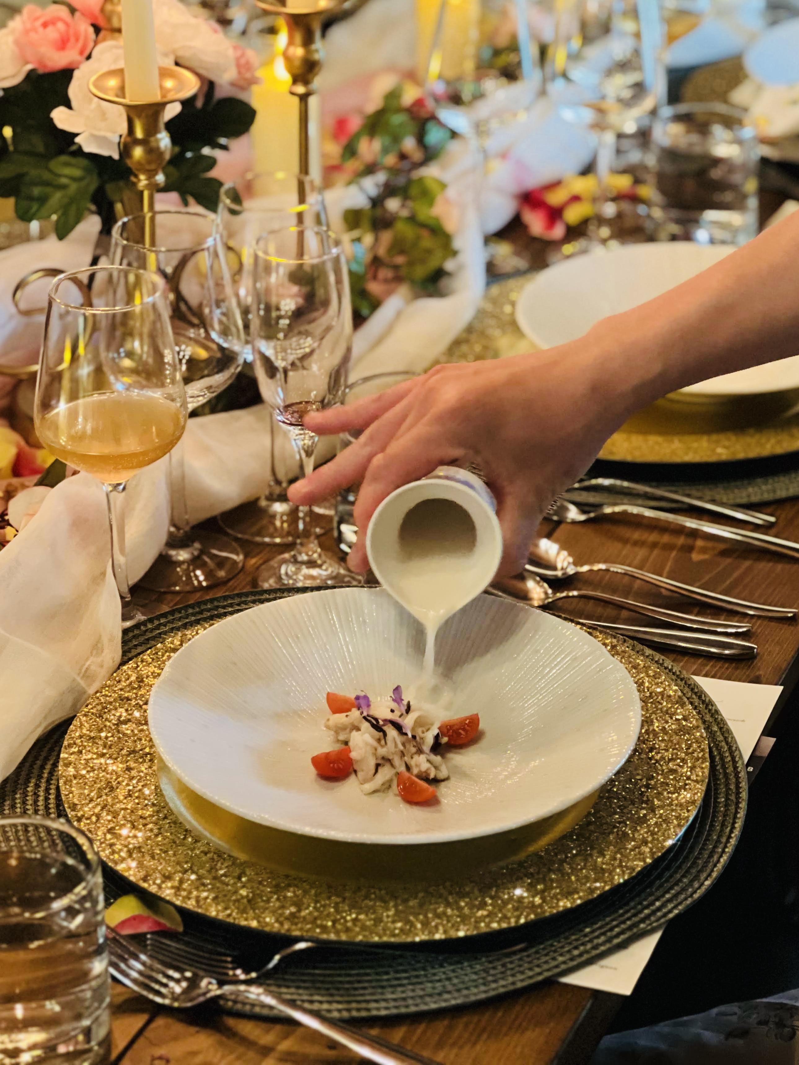 A hand pouring sauce tableside over a delicate first-course presentation