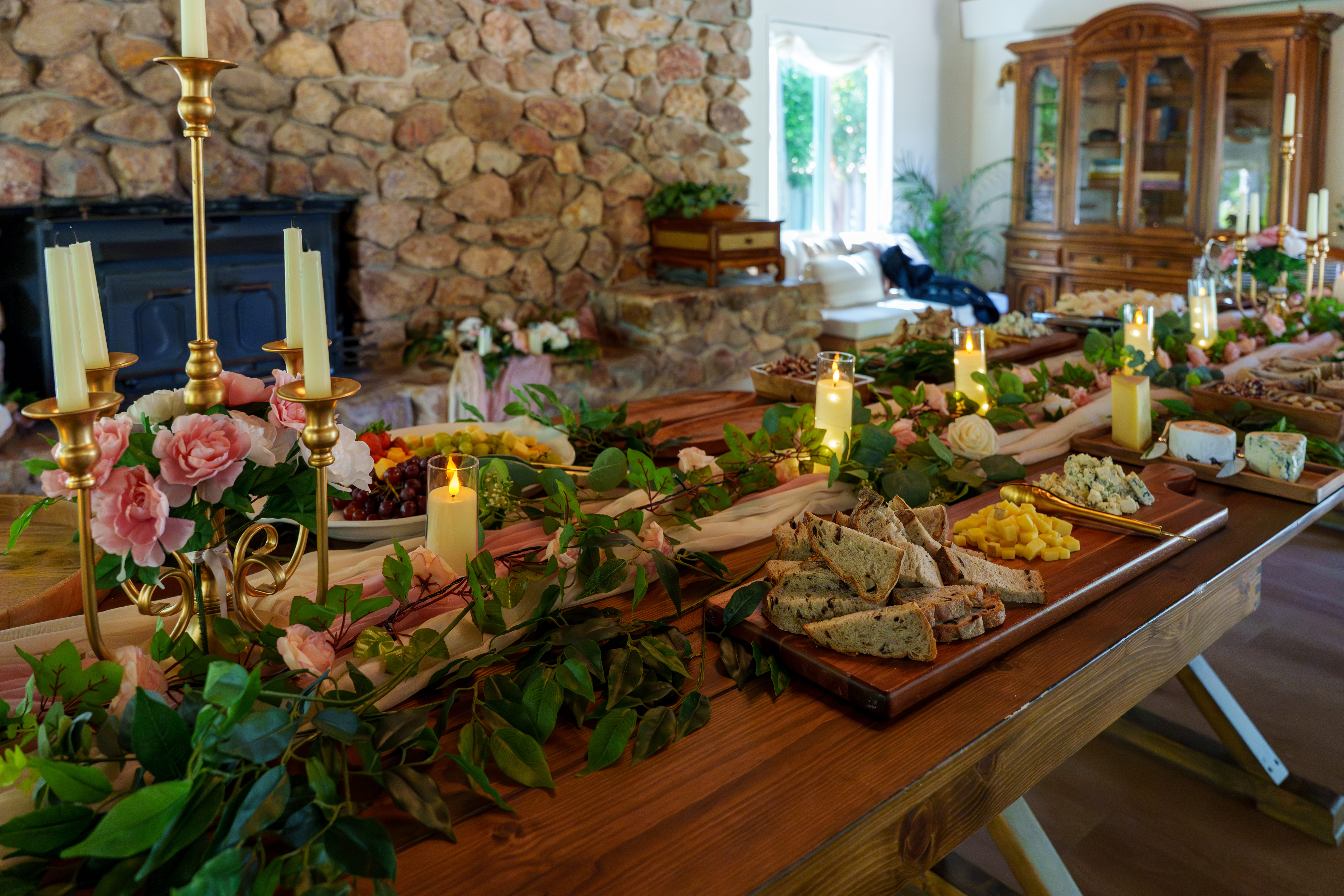 A long banquet table dressed with floral runners, candles, bread, and cheese boards by a stone hearth